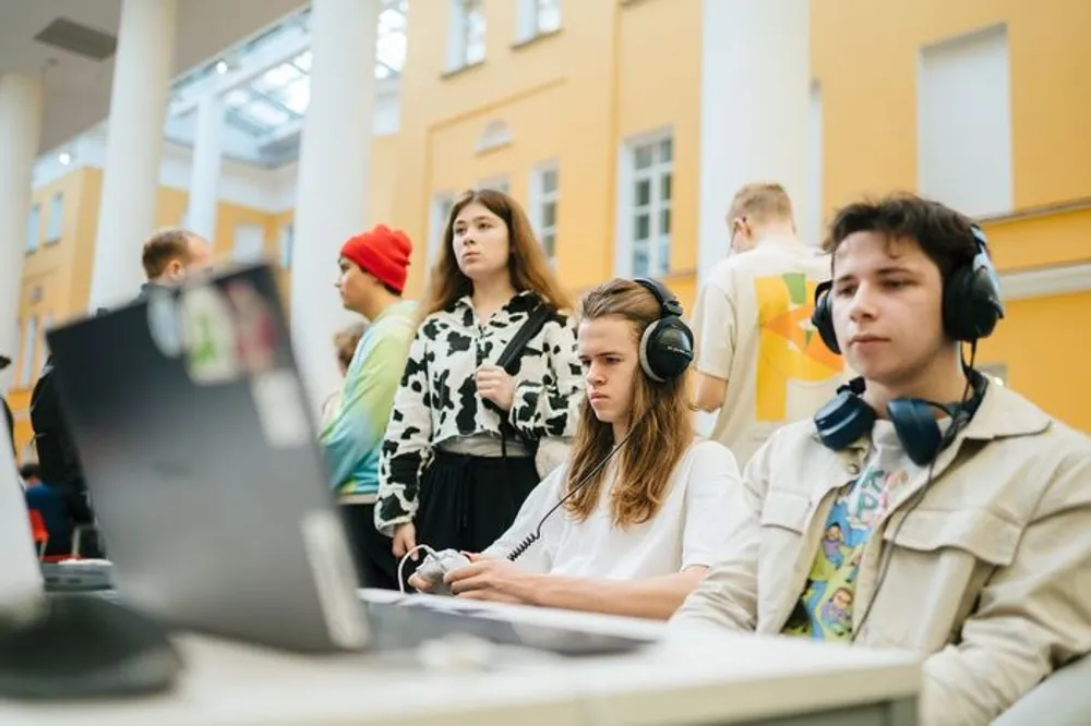 Группа молодых людей в помещении, занимающихся чем-то связанным с компьютерами. На переднем плане два человека с наушниками, остальные на заднем плане.