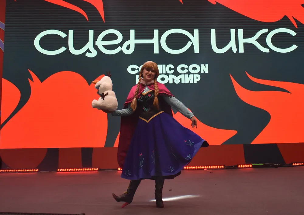 Изображение персонажа в костюме, возможно, с куклой, на фоне логотипа мероприятия. Косплей, сценическое выступление.