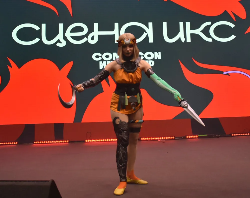 На изображении показан человек в костюме, вероятно, на мероприятии, связанном с комиксами или играми. Фон содержит текст, который может быть связан с названием мероприятия.