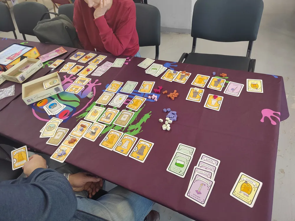 Изображение игрового стола с карточными играми и фигурками. На столе карты, фишки и коробки с играми.