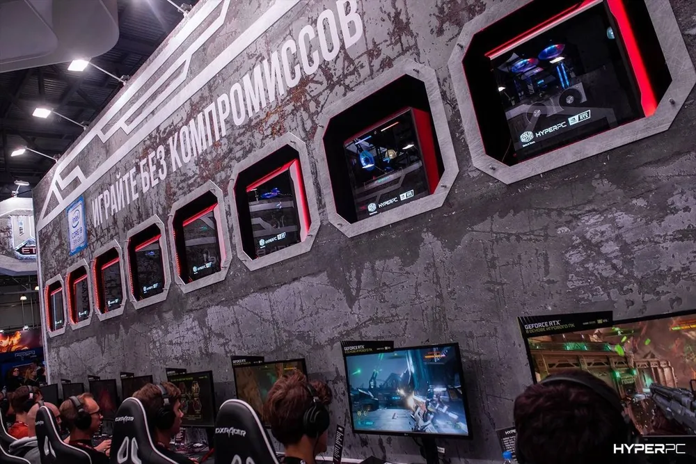 Изображение игрового стенда с компьютерами и экранами, на которых видны игровые сессии. Люди рядом с компьютерами, занимающиеся играми.