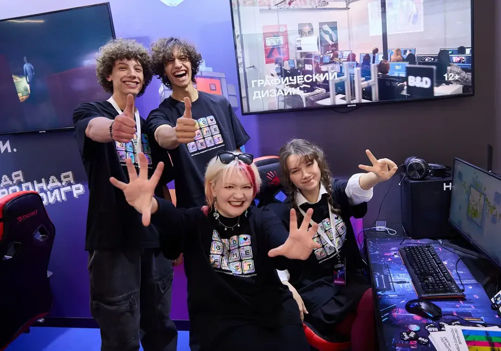 Четыре человека, стоящие на фоне компьютерной техники и экранов, позируют с улыбками и жестами. Обстановка связана с видеоиграми.