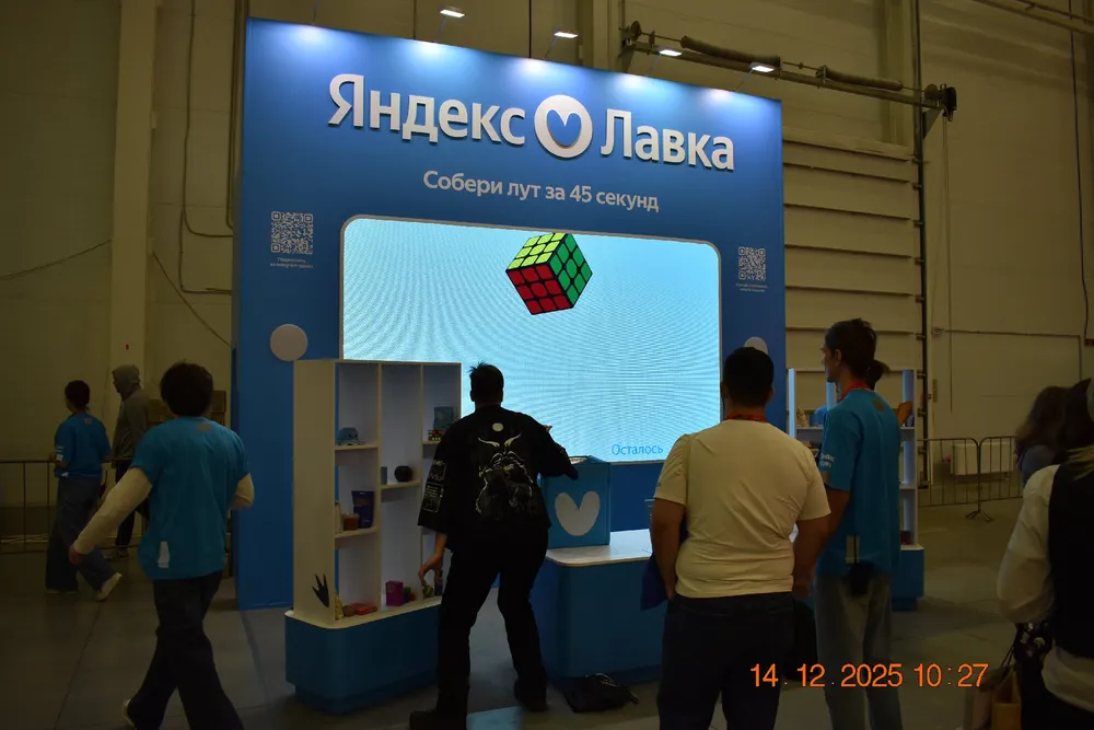 Изображение представляет собой стенд с логотипом Яндекса, на котором находится кубик Рубика и несколько людей в синей одежде.