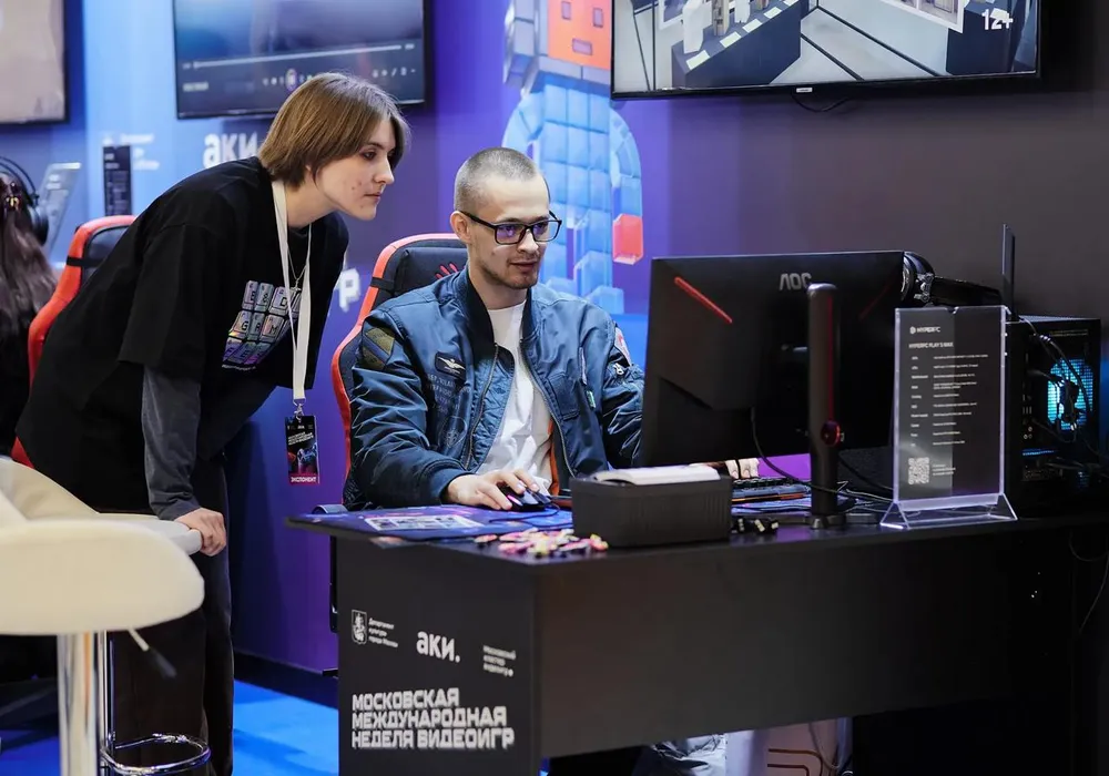 Изображение двух человек, один из которых сидит за компьютером, другой стоит рядом. Оба выглядят заинтересованными в процессе, обстановка современная.