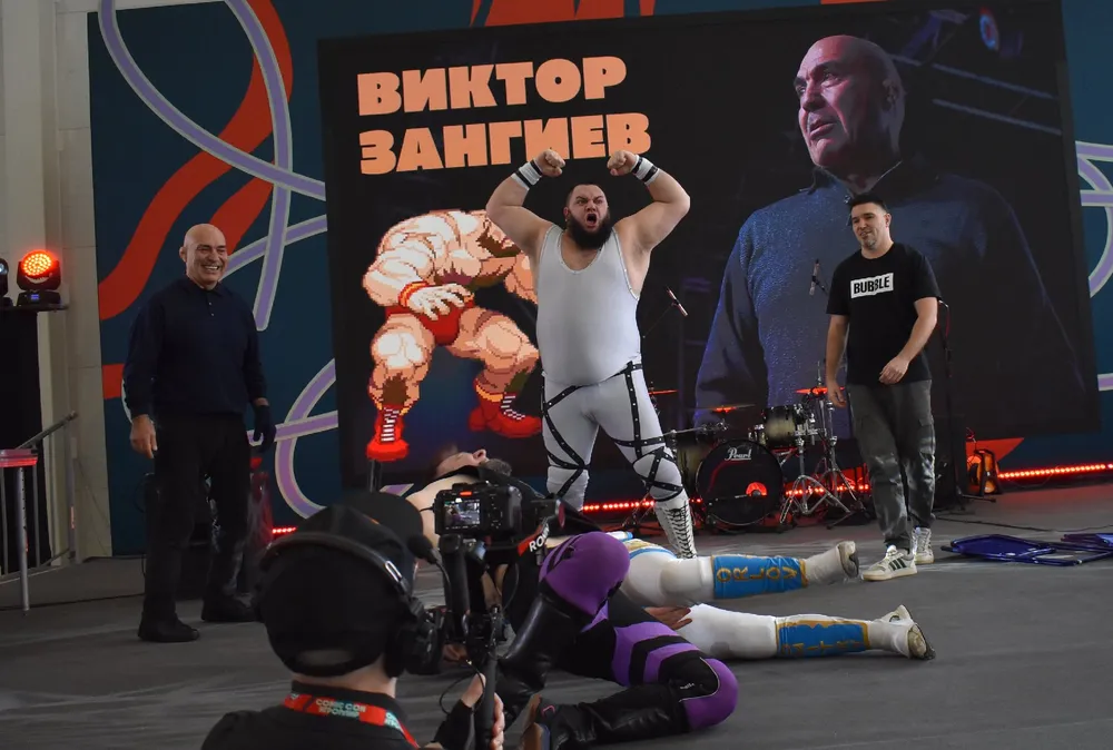 На изображении группа людей, один из которых в спортивной одежде, демонстрирует физическую силу на фоне большого баннера с текстом. Другие участники наблюдают.