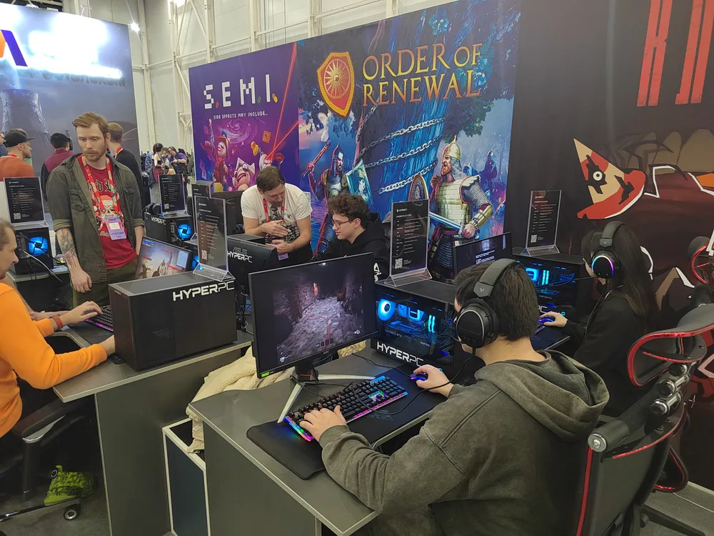 Сцена с несколькими людьми, играющими на компьютерах в игровом павильоне, с рекламой игр на стенде.