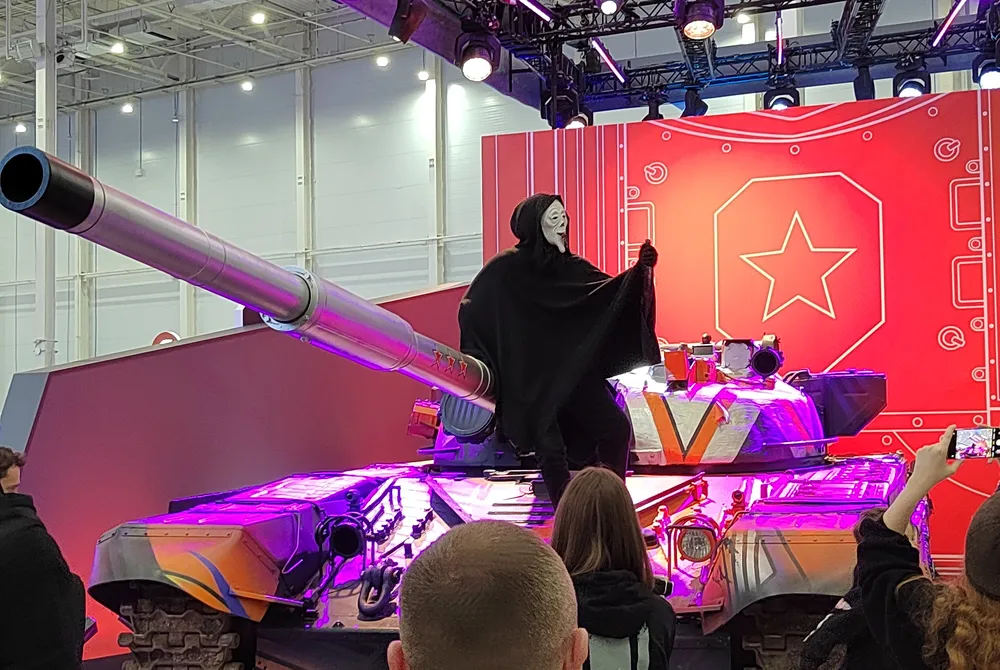 Изображение танка с фигурой в черной мантии на фоне красной стены. Видны люди, наблюдающие за сценой.