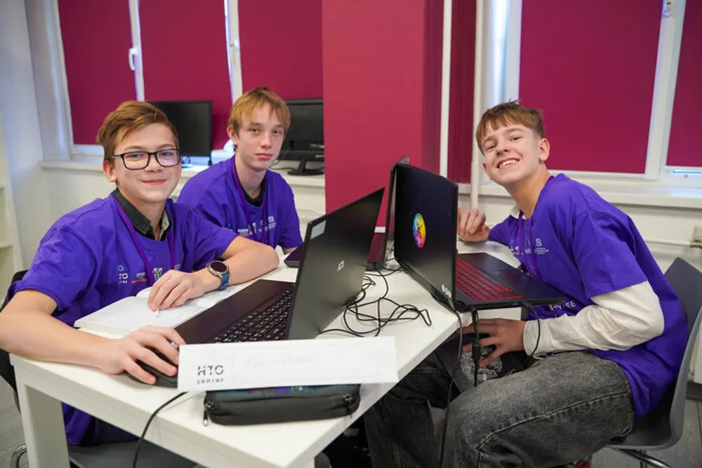Трое подростков за столом с ноутбуками, одеты в одинаковые фиолетовые футболки.