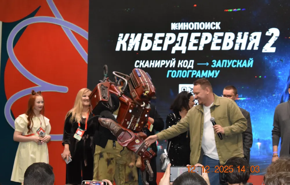 На изображении мероприятие, где люди на сцене, один из них в костюме. Фон с надписью \