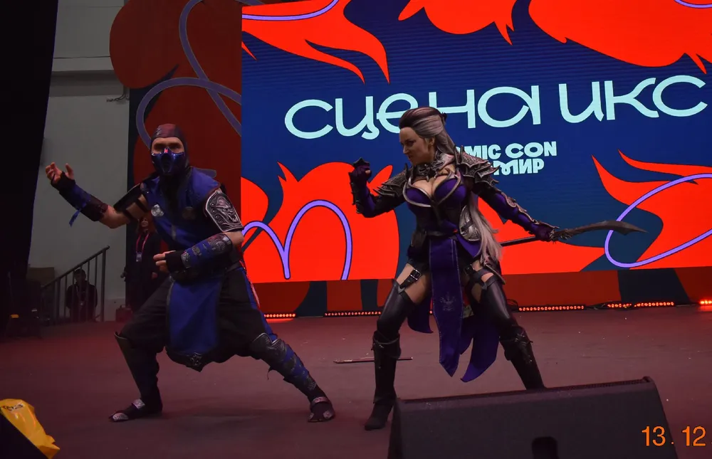 На изображении представлены люди в костюмах, выступающие на сцене. Сцена оформлена в стиле комиксов.