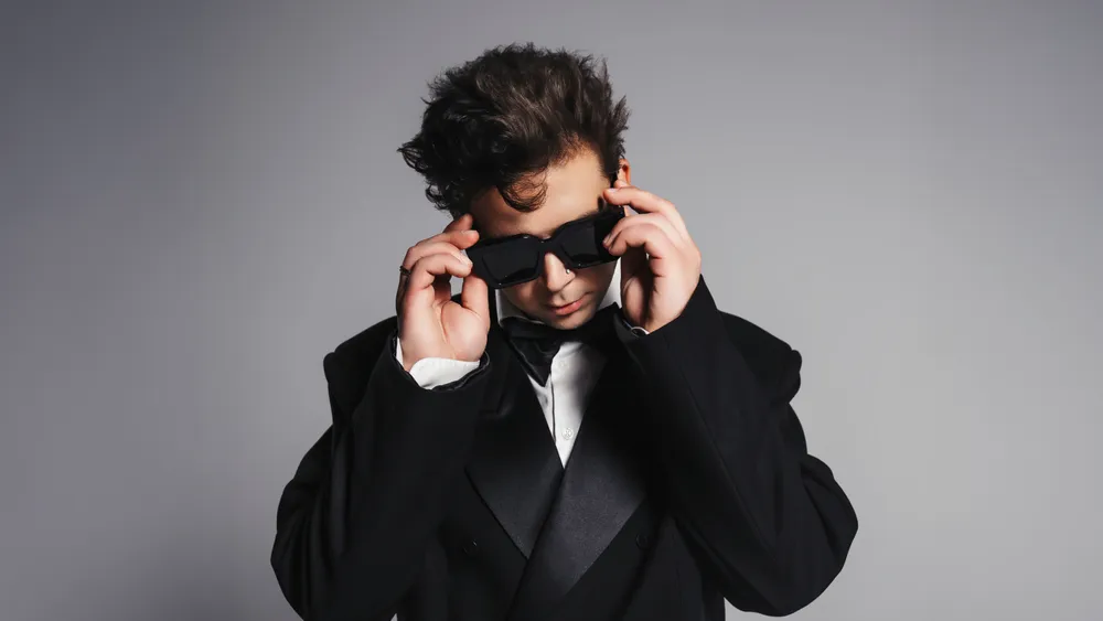 Молодой человек в черном костюме и темных очках, позирующий на фоне нейтральной серой стены.