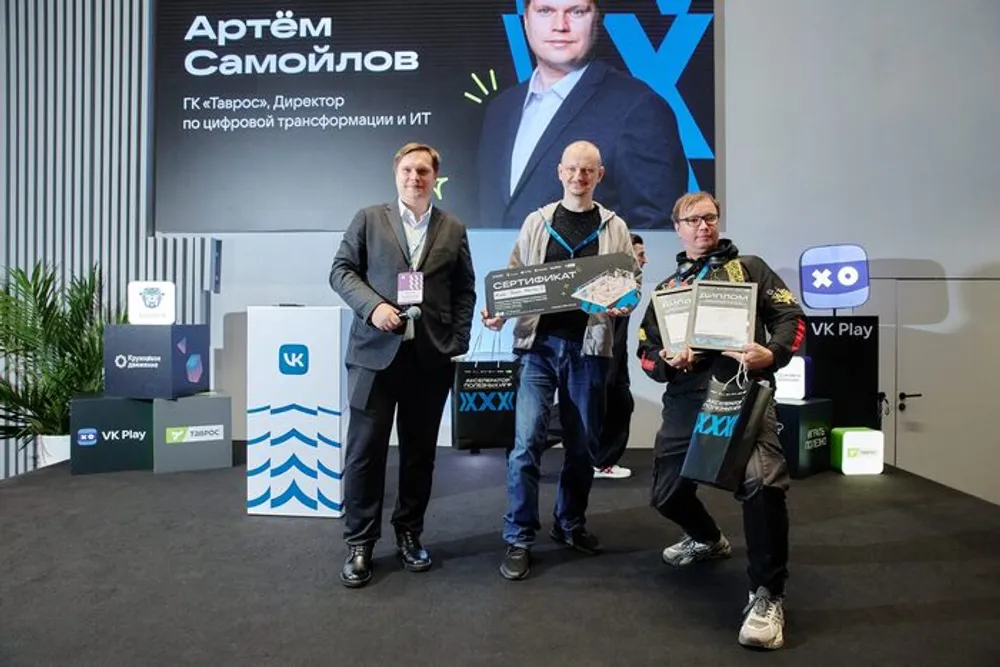 На изображении три человека на мероприятии, присутствуют награды и логотипы компаний.