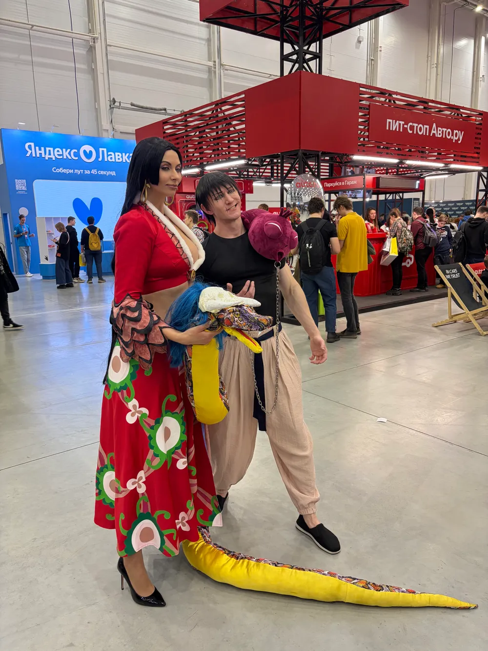 Две женщины в костюмах на мероприятии, одна в красном платье с цветами, другая в необычном наряде с яркими аксессуарами.