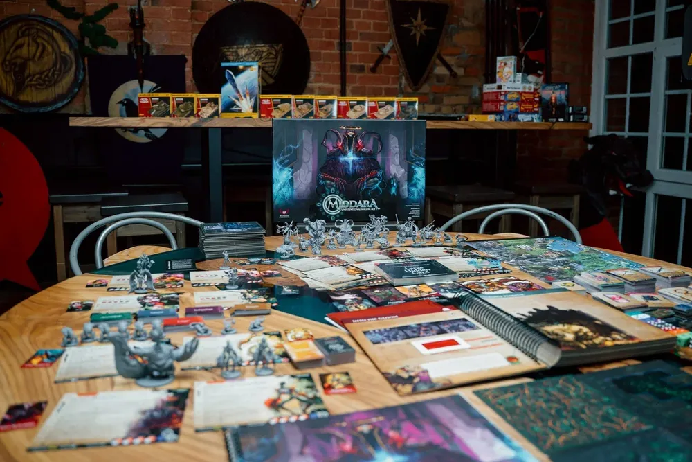 На изображении представлен стол с настольными играми и игровыми компонентами, включая миниатюры и карты.