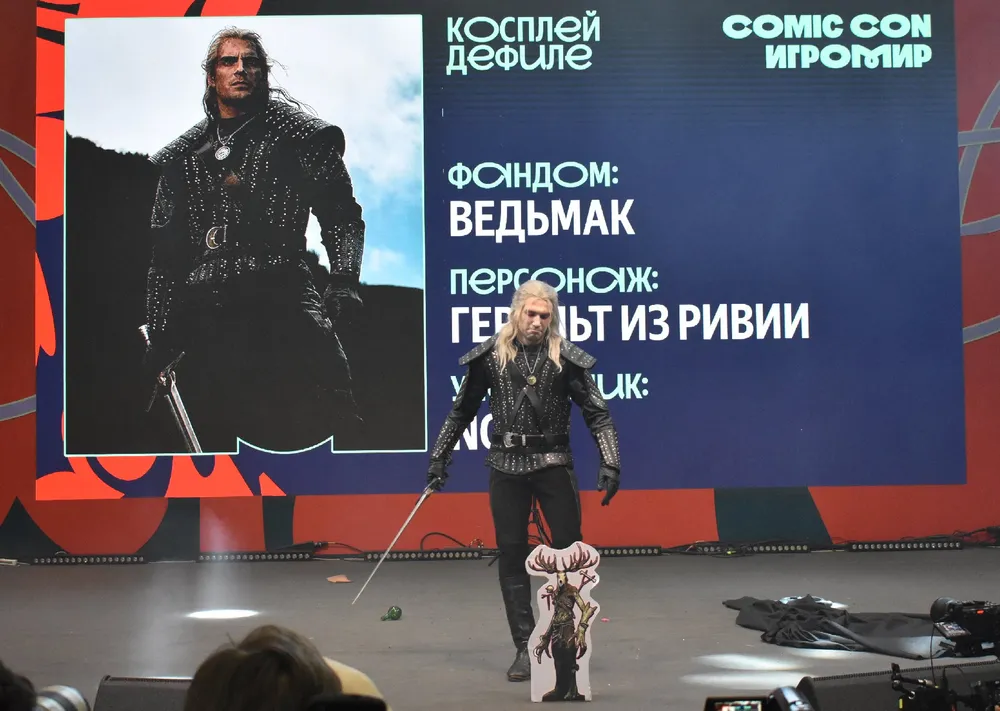 Изображение человека в костюме персонажа на мероприятии Comic Con, с изображением другого персонажа на экране.