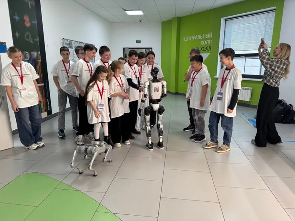 Группа детей и подростков в белых футболках, стоящих вокруг робота. В помещении с зелеными стенами и окнами.