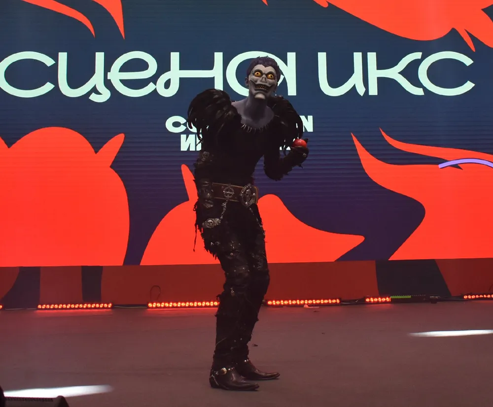 На изображении человек в костюме, выступающий на сцене. На заднем плане виден текст на экране.