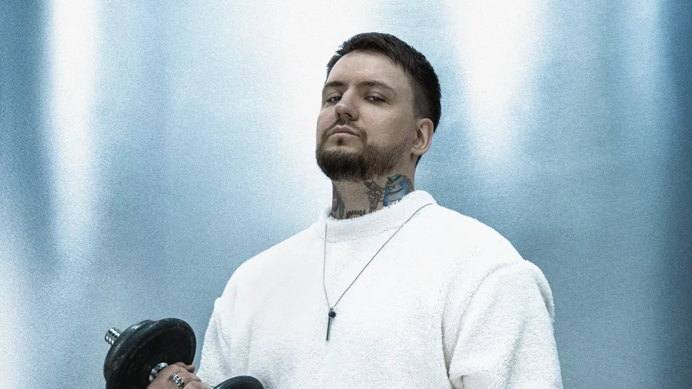 На картинке изображён человек с гантелями, в спортивной одежде, на фоне светлой стены.