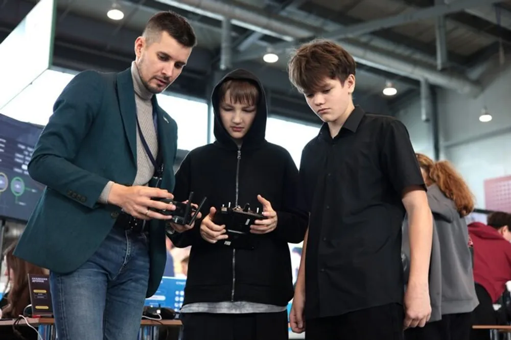 На изображении три человека, один из них объясняет что-то двум другим. Они держат устройства в руках и находятся в помещении с видимыми стендами.