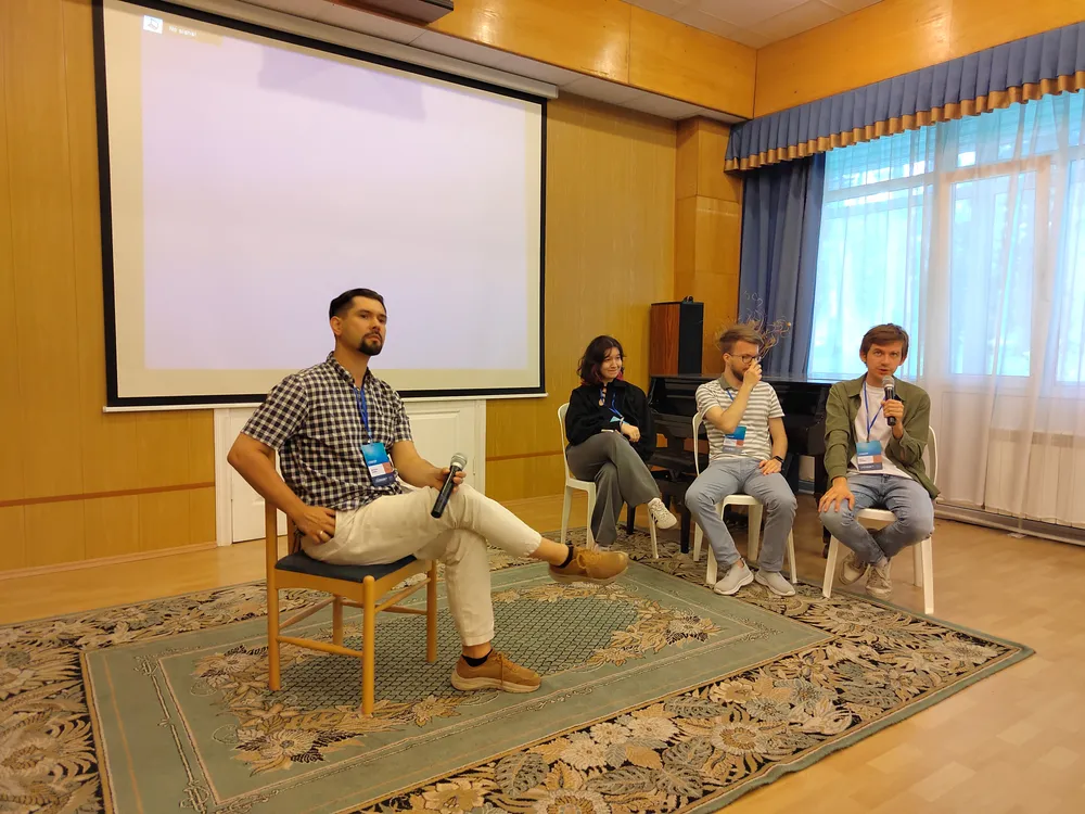 Четыре человека на сцене, один сидит на стуле, остальные стоят рядом. На заднем плане проектор и окно.