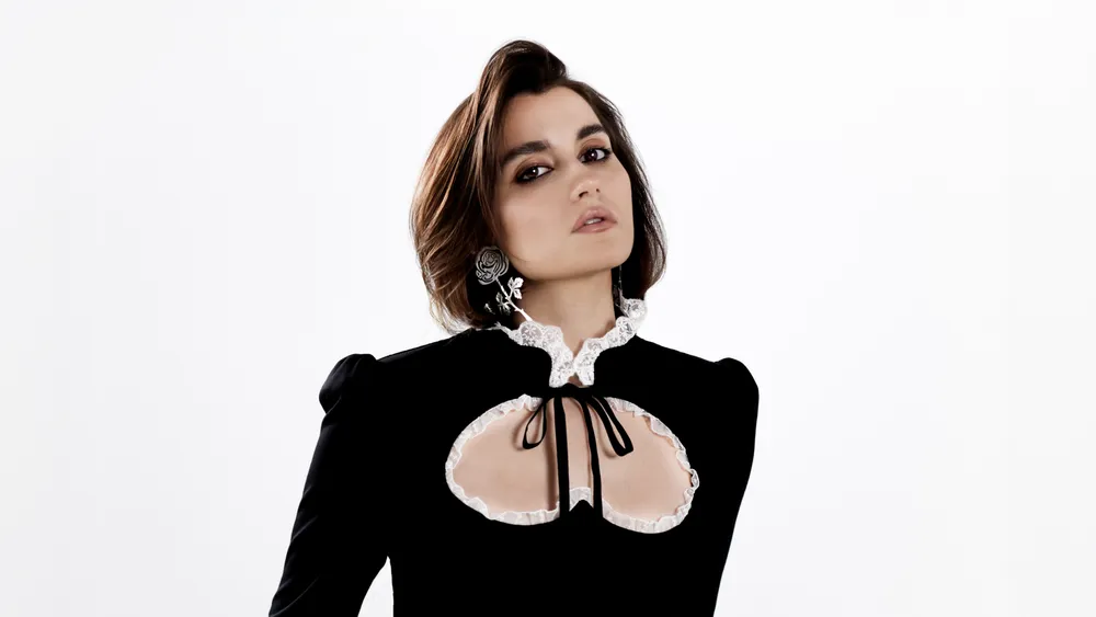 На картинке изображена женщина в платье с открытыми участками, позирующая на фоне белой стены.