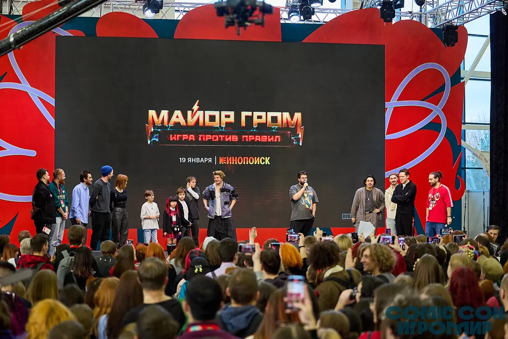 На изображении представлена сцена с людьми на мероприятии, возможно, связанном с игровой тематикой. Люди на сцене и в зале, экран с надписью \