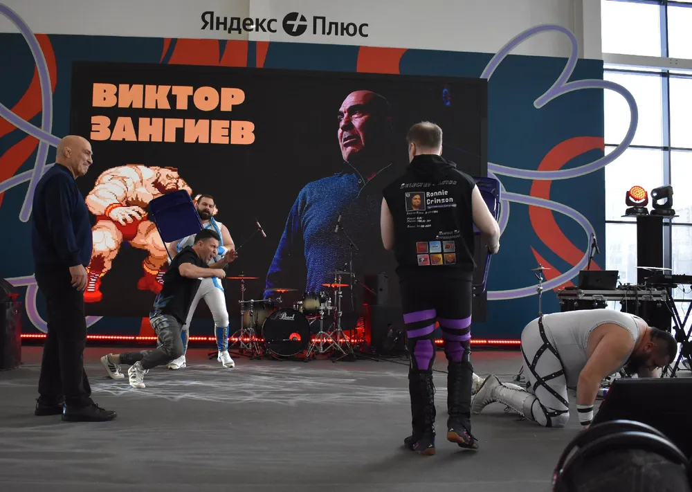 Сцена с несколькими людьми на мероприятии, на фоне экран с изображением. Люди в спортивной одежде, возможно, занимаются борьбой или демонстрацией.