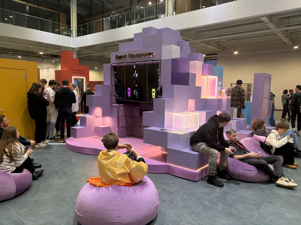 Изображение показывает людей в помещении, многие из которых сидят на мягких пуфах. В центре находится структура, напоминающая игровую зону с экраном. Атмосфера дружелюбная и непринужденная.