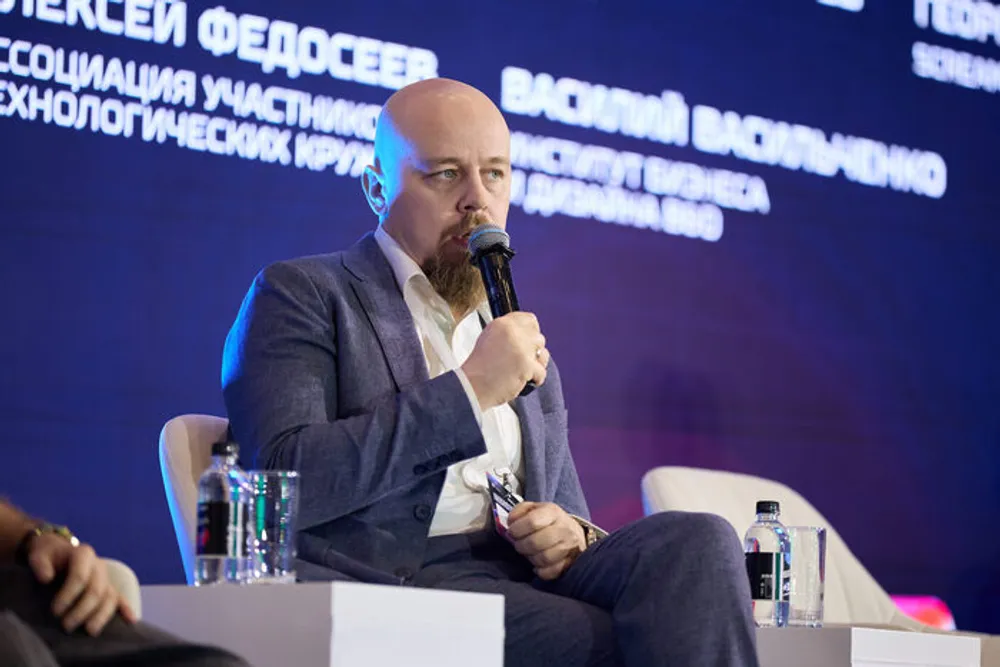 На изображении мужчина на мероприятии, вероятно, выступает или участвует в обсуждении. Видно, что он держит микрофон.