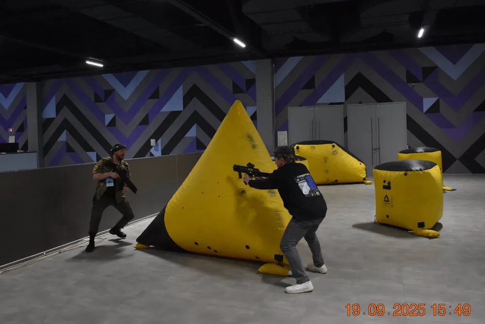 Два человека, одетые в спортивную одежду, играют в активную игру в закрытом помещении. Вокруг них расположены желтые конструкции, напоминающие укрытия. Фон состоит из геометрических узоров.