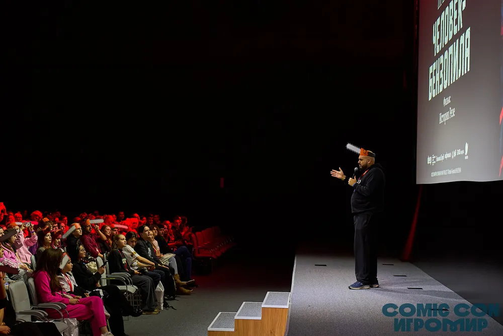 Сцена с выступающим человеком на сцене перед аудиторией в зале. На заднем плане виден экран с текстом.