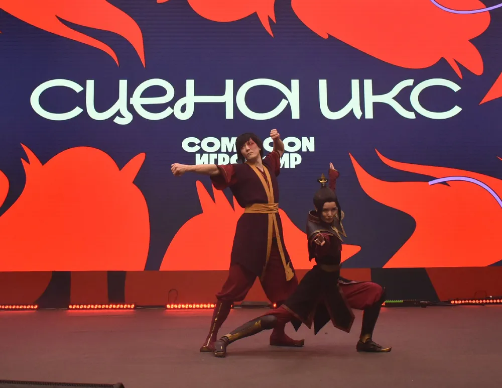 Изображение людей в костюмах, выступающих на сцене. На заднем плане видно название мероприятия.