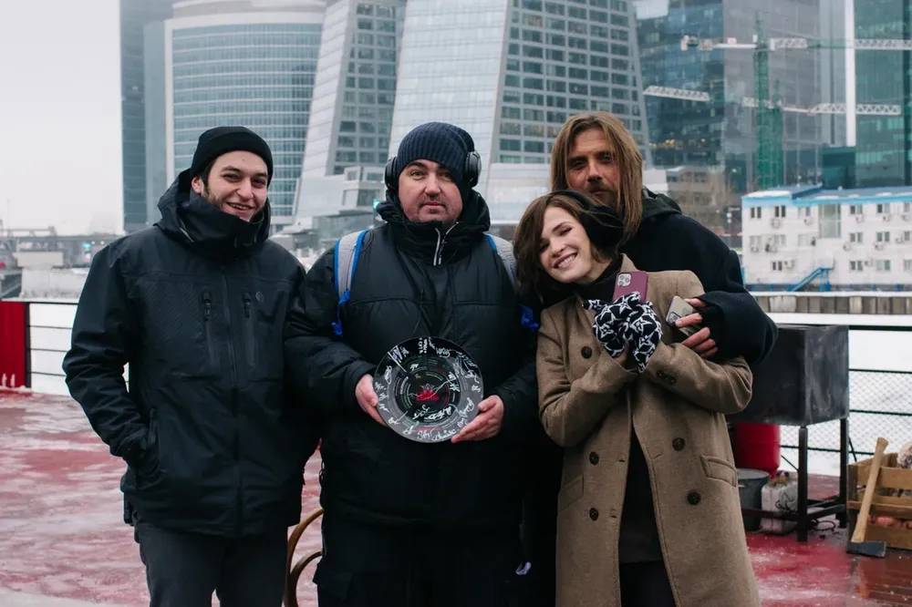 Группа людей на открытом воздухе, стоят на фоне зданий. Один из мужчин держит приз или награду, остальные улыбаются.