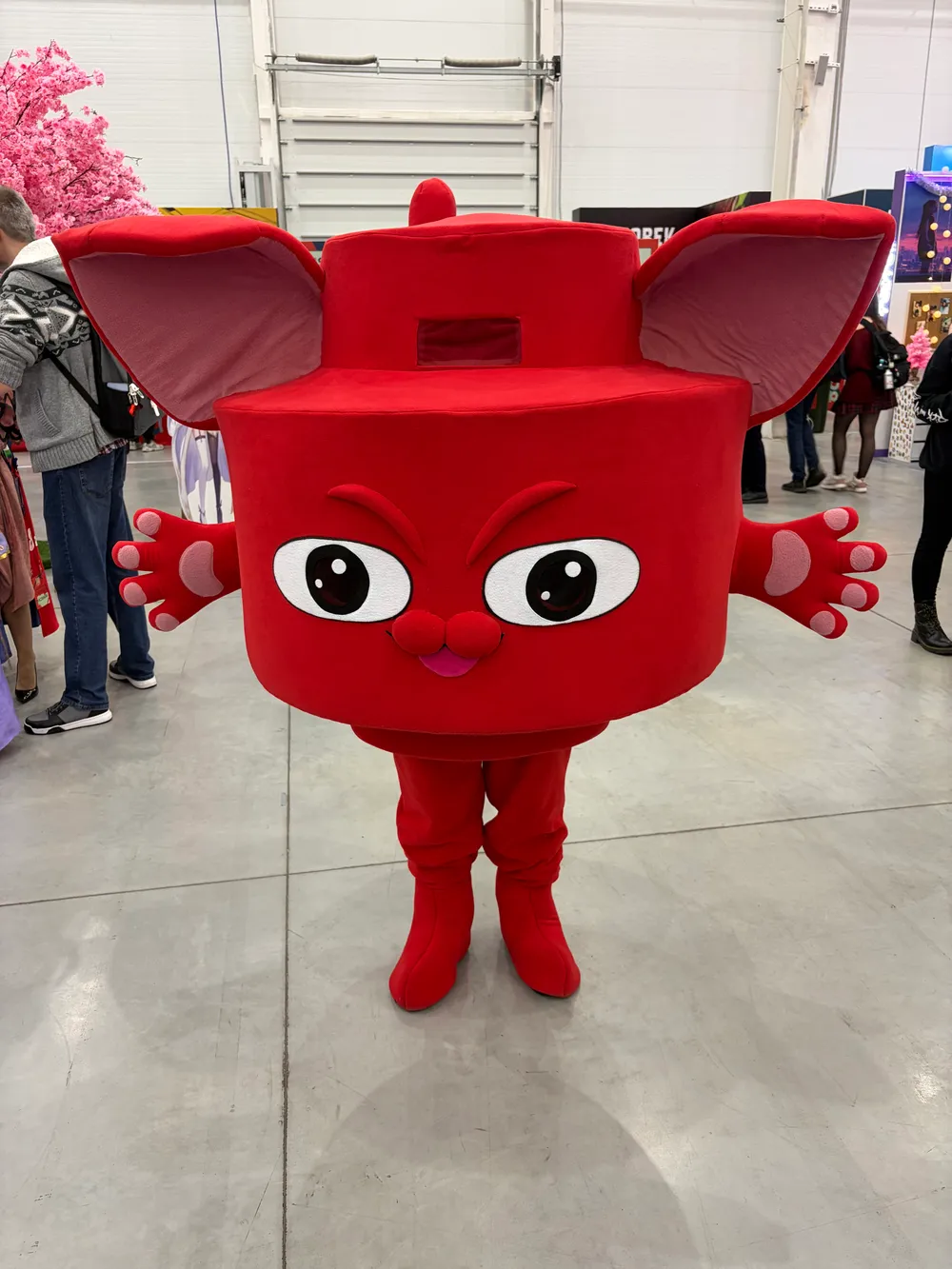 На изображении представлен персонаж в костюме, напоминающем красную игрушку с большими глазами и ушами, стоящий на выставке.