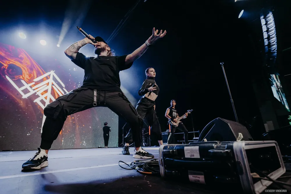 На изображении группа музыкантов выступает на сцене, один из них держит микрофон и активно исполняет песню. Сцена ярко освещена, на заднем плане виден световой экран.