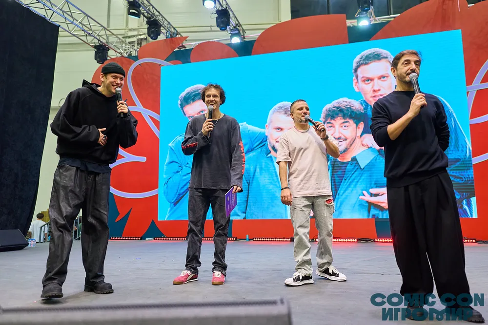 Четыре человека на сцене, выступающие с микрофонами, фон с ярким экраном.