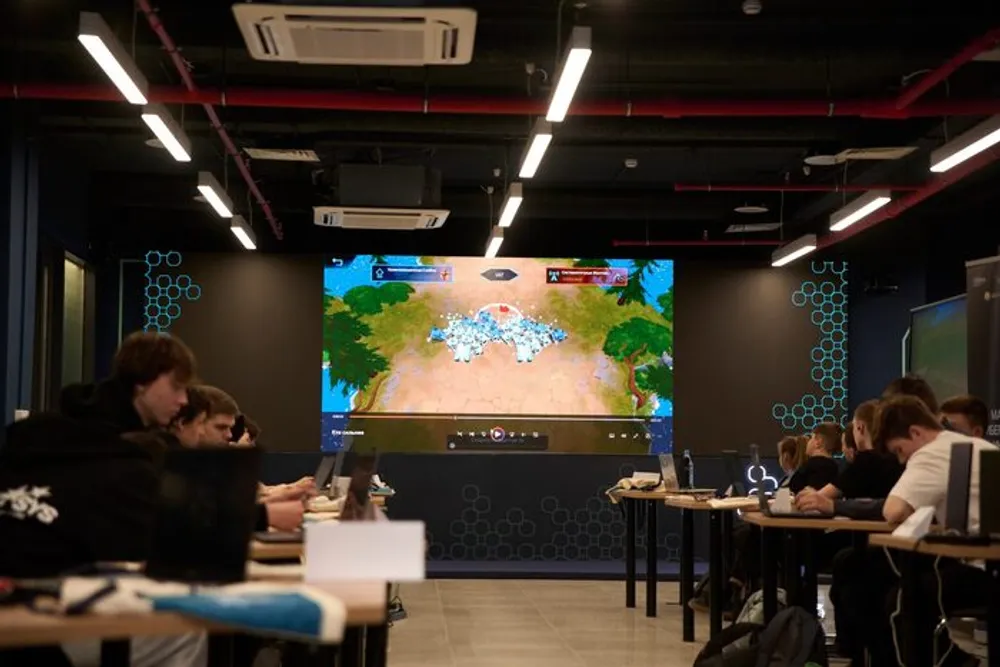 На изображении показан класс с людьми, которые работают за столами, и большой экран, на котором отображается игровая сцена.