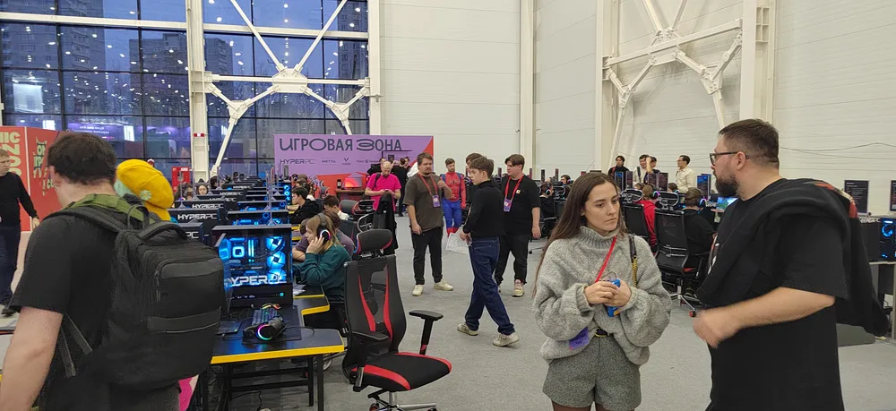 Изображение людей на мероприятии, возможно, выставка или конференция, с компьютерами и игровыми креслами.