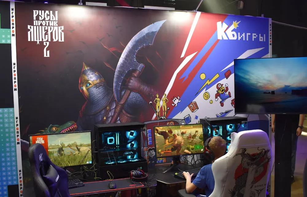 Изображение игрового стенда с компьютерами, на которых запущены игры. На заднем плане рекламный баннер с изображением военной тематики.