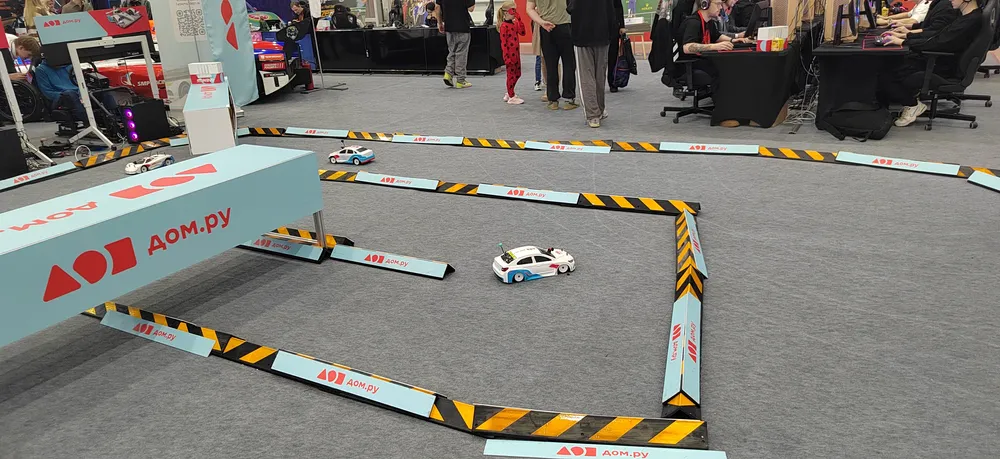 На изображении представлена трасса для гонок на радиоуправляемых автомобилях, с участниками и зрителями на фоне.