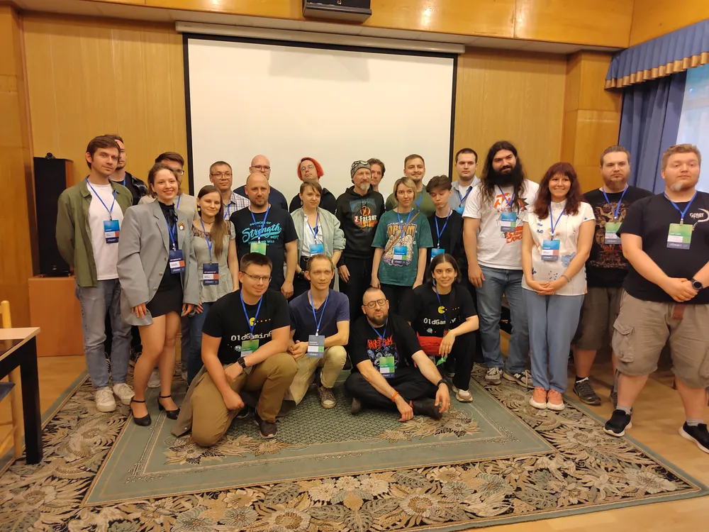 Группа людей на мероприятии, некоторые в футболках, все улыбаются, на заднем плане проектор и деревянные стены.