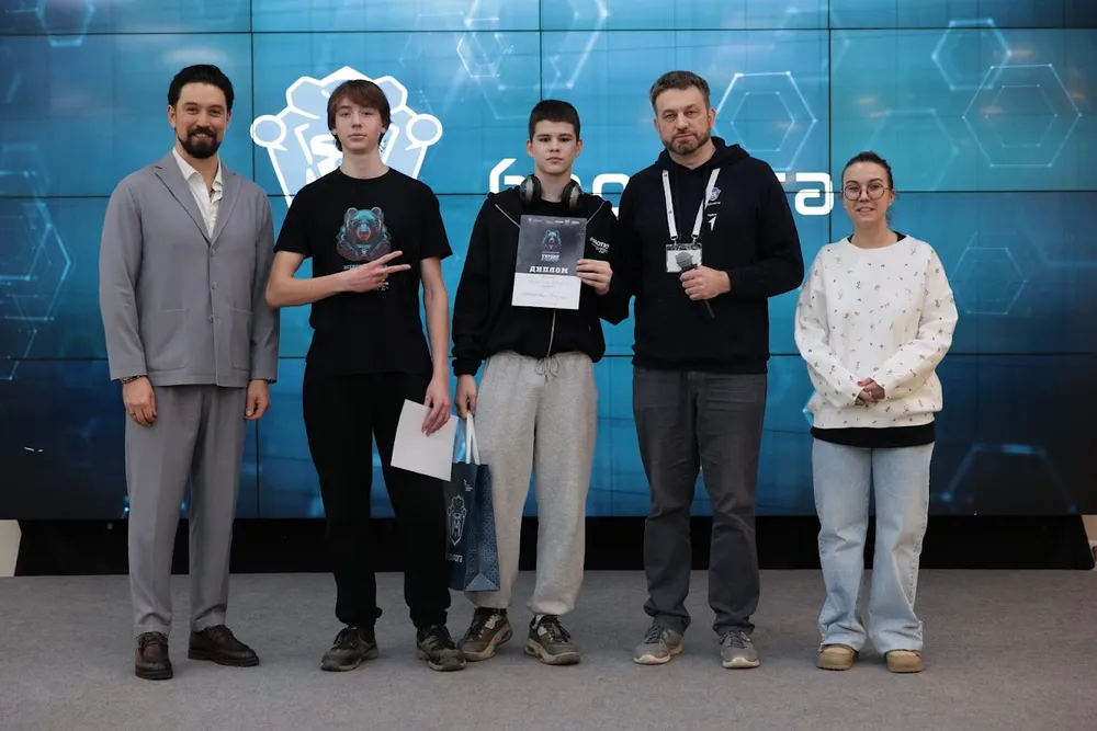 Группа людей на мероприятии, вероятно, награждение или церемония. Четыре человека стоят на фоне технологической стены, один из них держит документ.