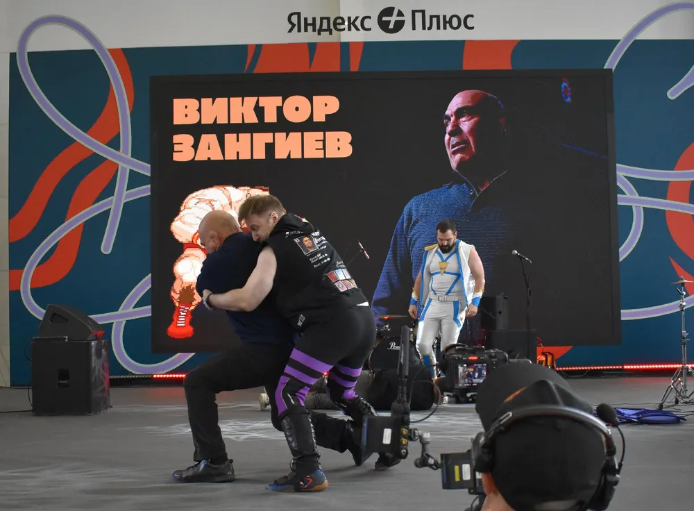 На картинке изображены люди на мероприятии, один из них выполняет физическое упражнение, на фоне виден экран с текстом и изображением.