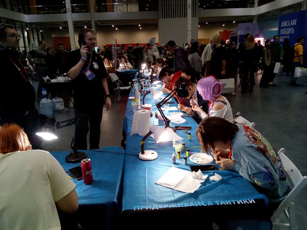 Изображение людей, занимающихся деятельностью за столами, возможно, в рамках мероприятия или мастер-класса. На заднем плане видно людей и оборудование.