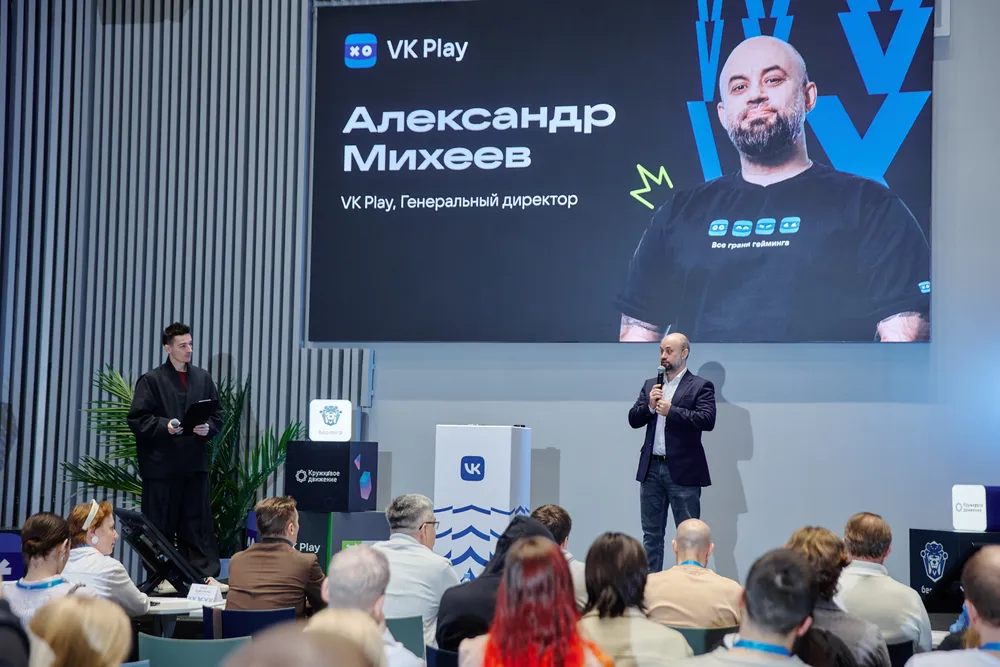 На изображении видно выступление человека на сцене, за ним находится экран с текстом, на котором указано имя и должность. В зале присутствует аудитория.