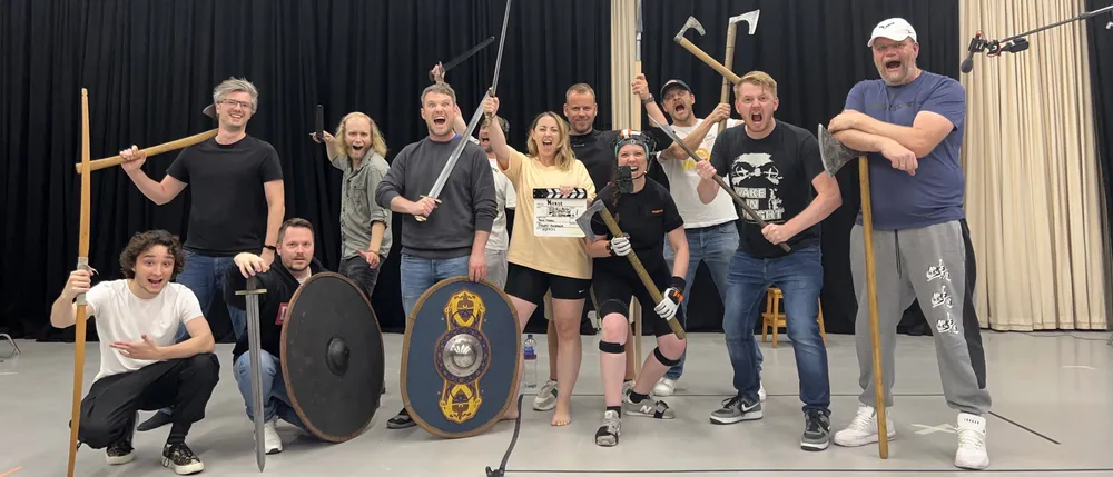 Группа людей позирует на фоне сцены, некоторые держат оружие, такие как копья и щиты. Внешний вид участников разнообразен.