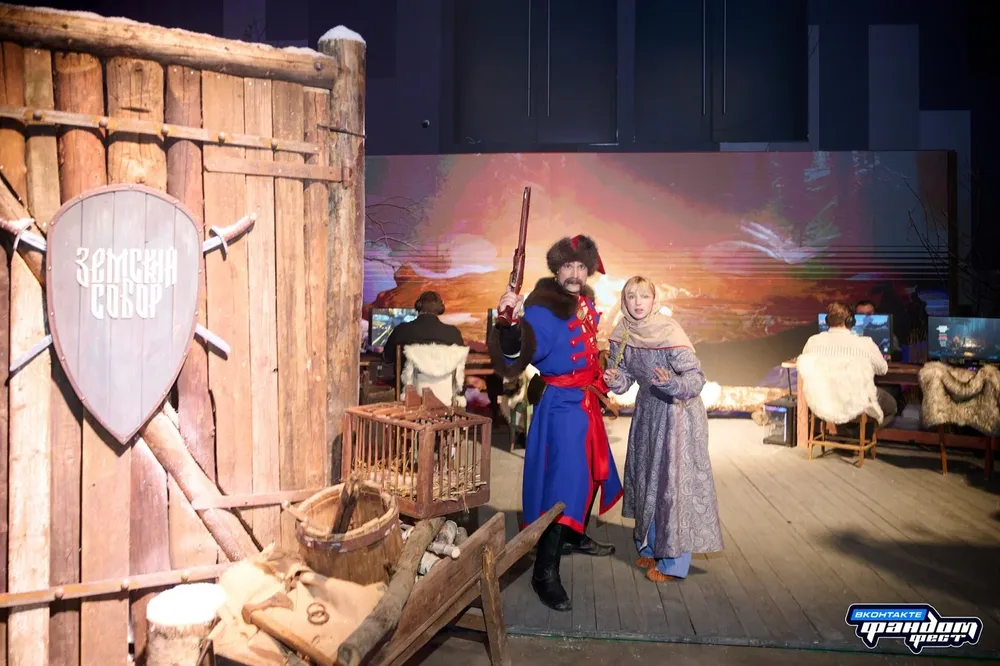 На изображении сцена с персонажами в исторической или костюмированной обстановке. Один человек в костюме с оружием, другой в исторической одежде. Фон с деревянной конструкцией и декорациями, напоминающими старинный стиль.