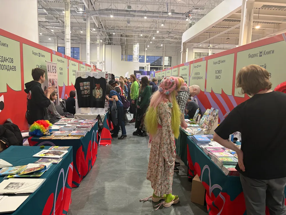 Изображение показывает людей на мероприятии с выставочными стендами, некоторые участники одеты в яркие костюмы, присутствуют столы с товарами.
