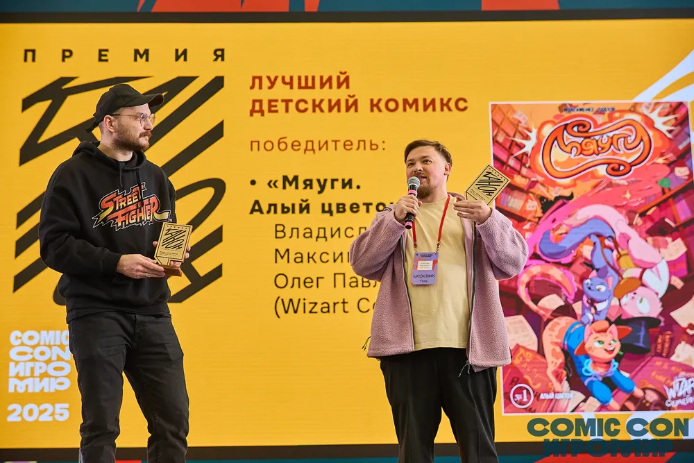 На изображении два человека на сцене, один из них держит награду, а другой говорит. Позади них находится баннер с текстом о премии и обложкой комикса.