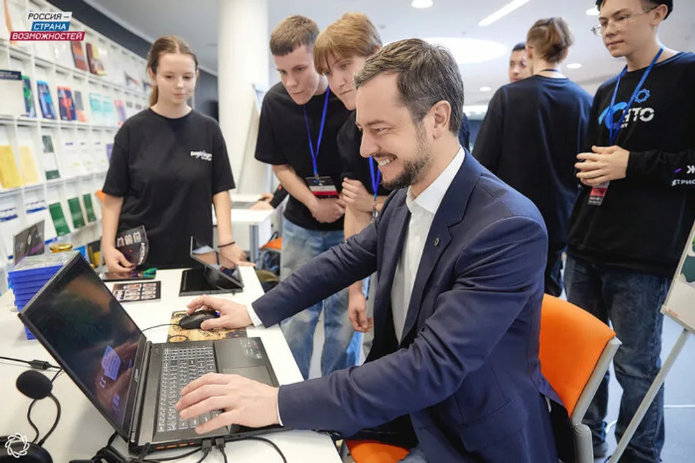 На изображении группа людей, наблюдающих за мужчиной, работающим за ноутбуком, на фоне стенда с информационными материалами.